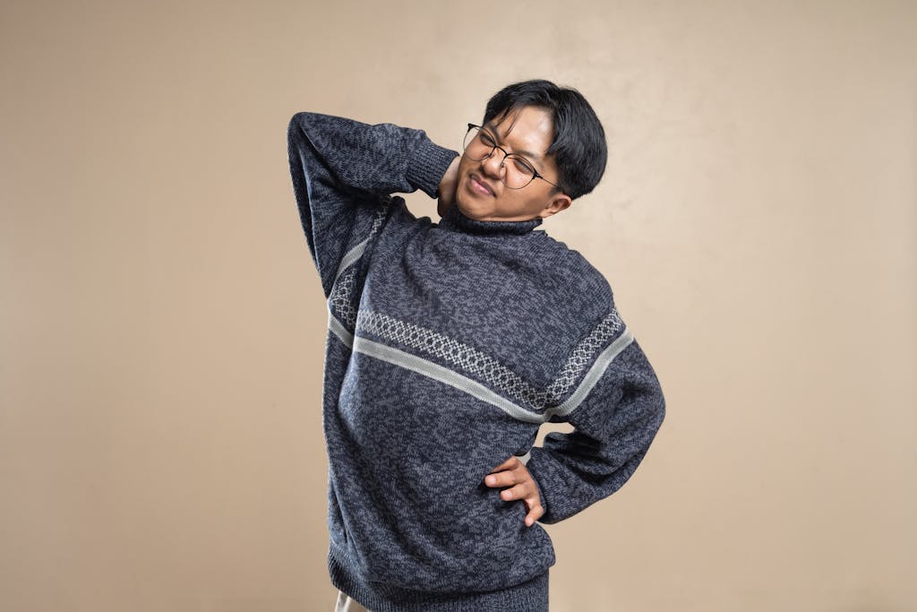 Asian man wearing eyeglasses and sweater stretching indoors, feeling body pain.