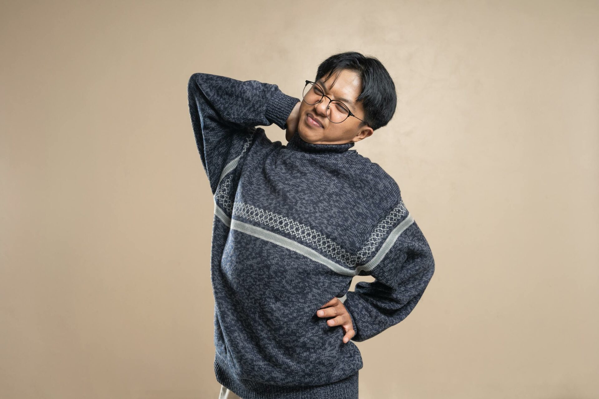 Asian man wearing eyeglasses and sweater stretching indoors, feeling body pain.